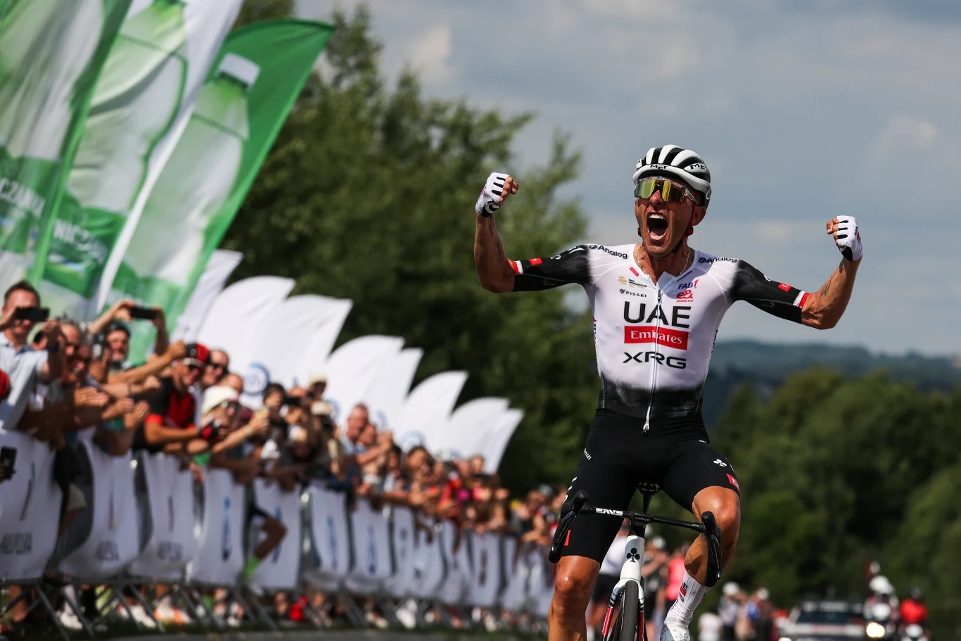 kolarz w stroju drużyny UAE cieszy się na mecie wyścigu rowerowego, unosi ręce w geście zwycięstwa, wokół tłum kibiców z flagami i zielonymi banerami, w tle zielone drzewa