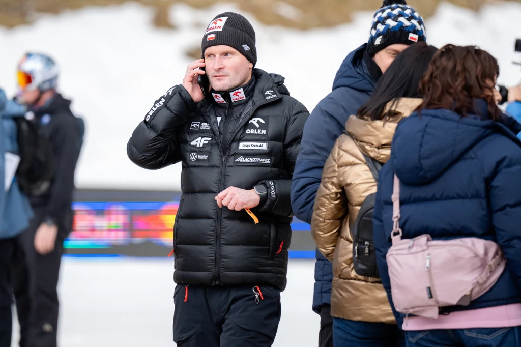 Mężczyzna ubrany w zimową kurtkę i czapkę z logo sponsorów rozmawia przez telefon, otoczony przez innych ludzi w zimowej scenerii, na tle rozmytego krajobrazu z elementami śniegu.