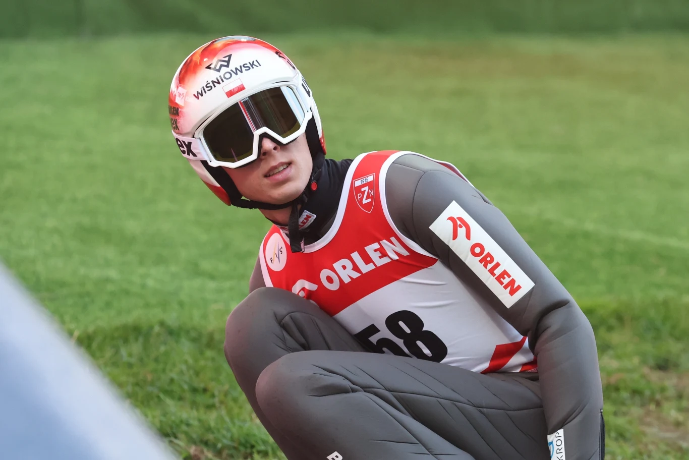 Zawodnik w kombinezonie narciarskim i kasku, oznaczony numerem 58, siedzi na trawie podczas zawodów skoków narciarskich; na kasku i stroju widoczne są logotypy sponsorów.