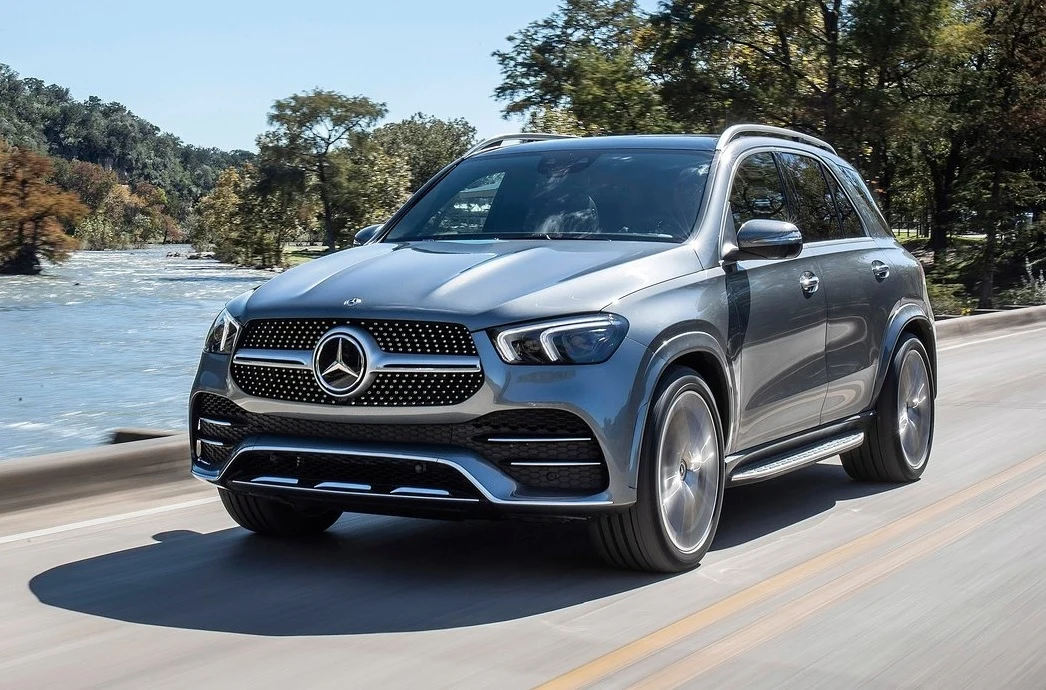 Srebrny samochód SUV marki Mercedes-Benz jedzie wzdłuż drogi nad rzeką, otoczony zielenią i drzewami, pojazd dominuje kadr, a tło pozostaje lekko rozmazane przez ruch.