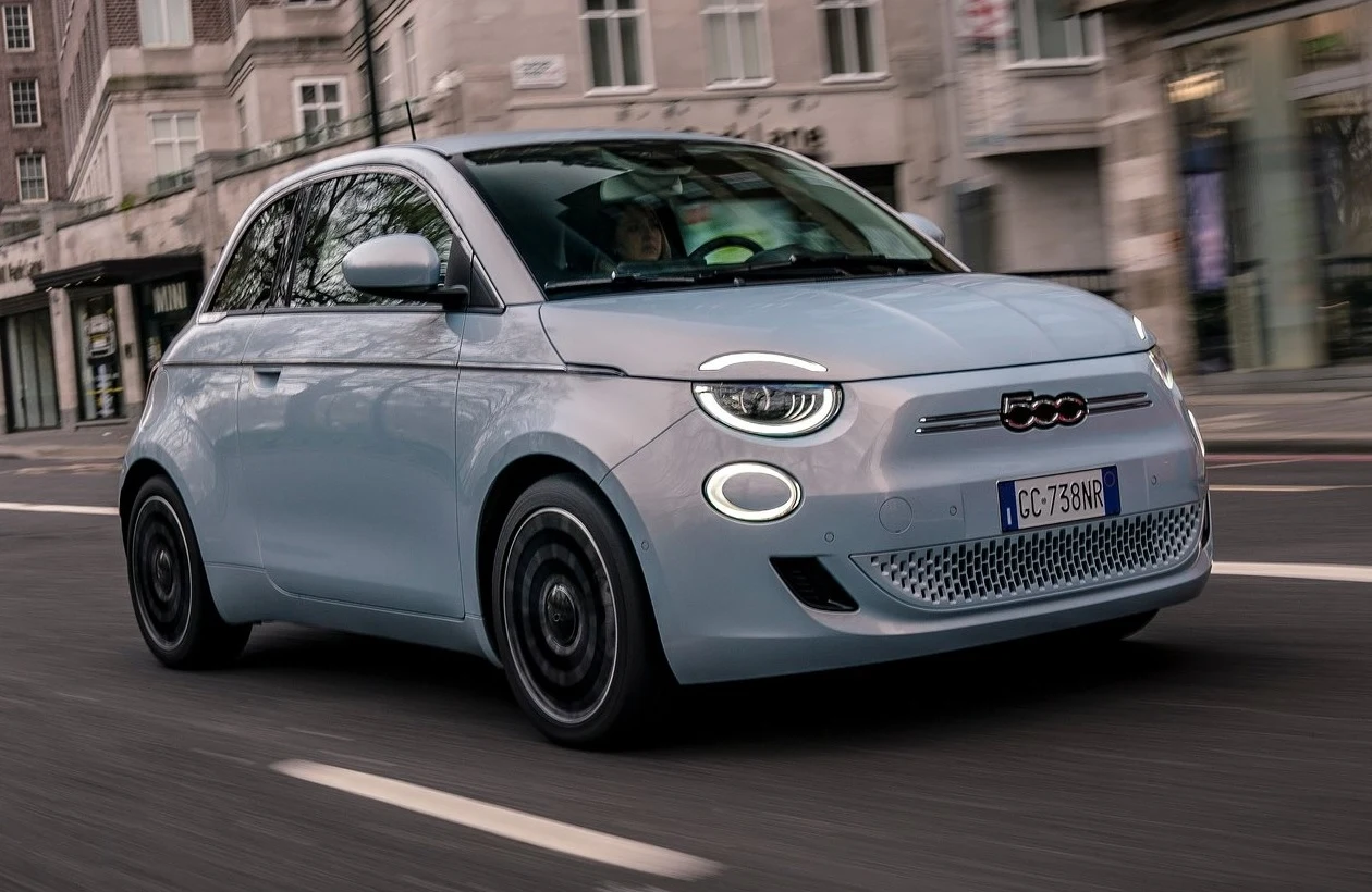 Jasnoniebieski samochód elektryczny Fiat 500 poruszający się po ulicy miejskiej z zabudową w tle, charakterystyczne okrągłe światła LED na froncie oraz włoska tablica rejestracyjna.