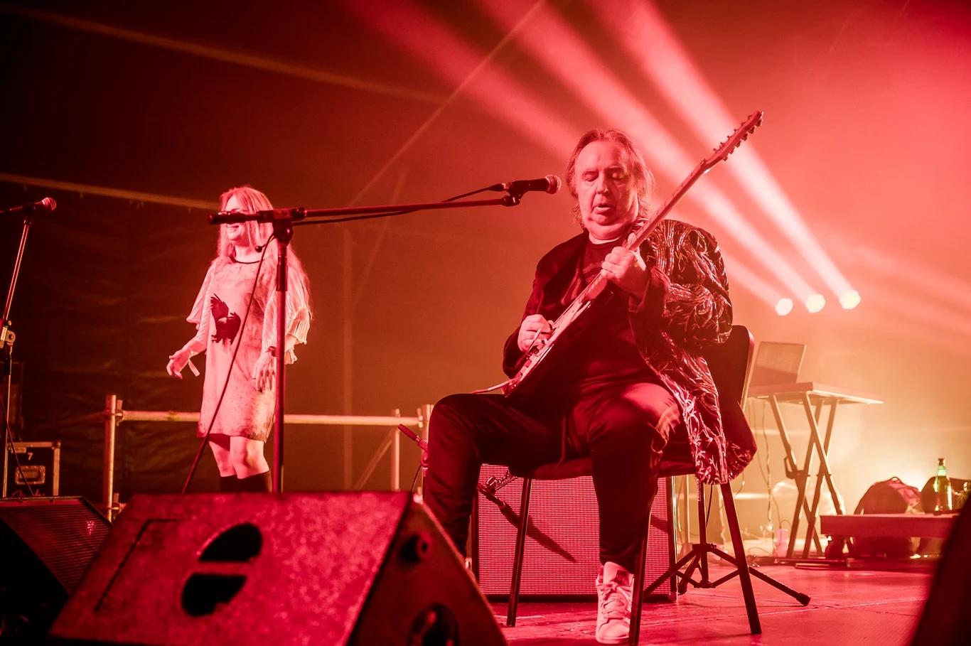 Mężczyzna siedzący na krześle gra na gitarze elektrycznej podczas koncertu, w tle kobieta śpiewająca przy mikrofonie, całość oświetlona intensywnym czerwonym światłem sceny.