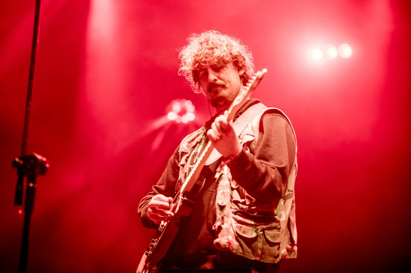 Muzyk z kręconymi włosami grający na gitarze elektrycznej w czerwonym oświetleniu scenicznym, dynamiczny klimat koncertu, wyraźny fokus na artystę i jego instrument.