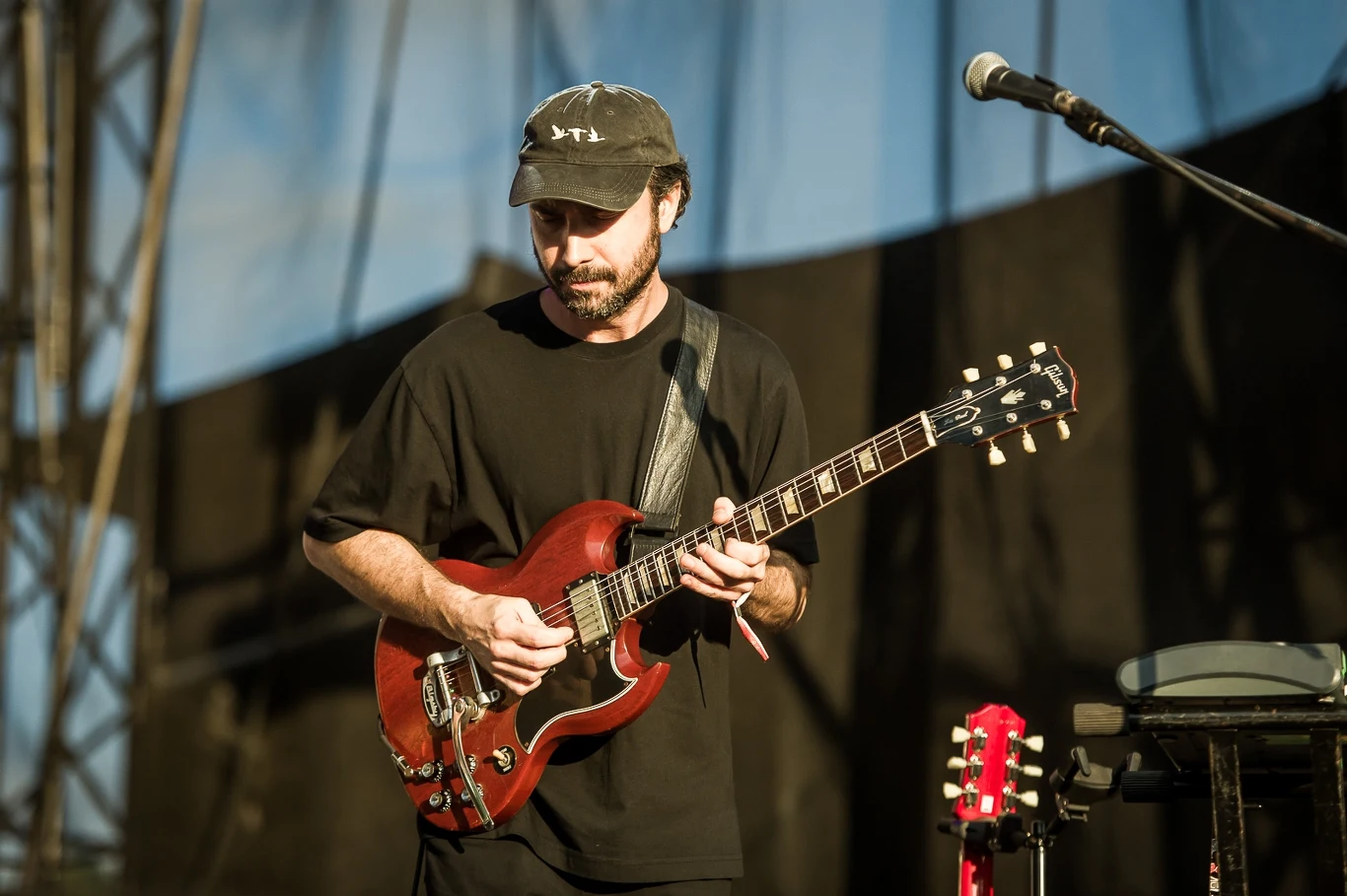 Mężczyzna w czarnej koszulce i czapce grający na czerwonej gitarze elektrycznej na scenie, z mikrofonem ustawionym z boku, w tle elementy sceniczne i niebieskie światło.