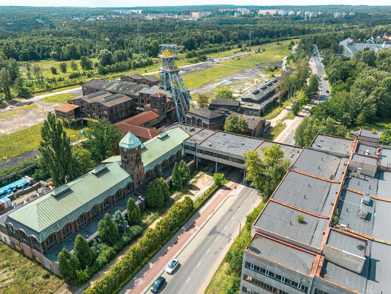 Kompleks budynków przemysłowych z wieżą szybową kopalni, otoczony zielenią i drogą prowadzącą przez środek kadru, w tle widać rozległe tereny zielone oraz dalszą zabudowę miejską.