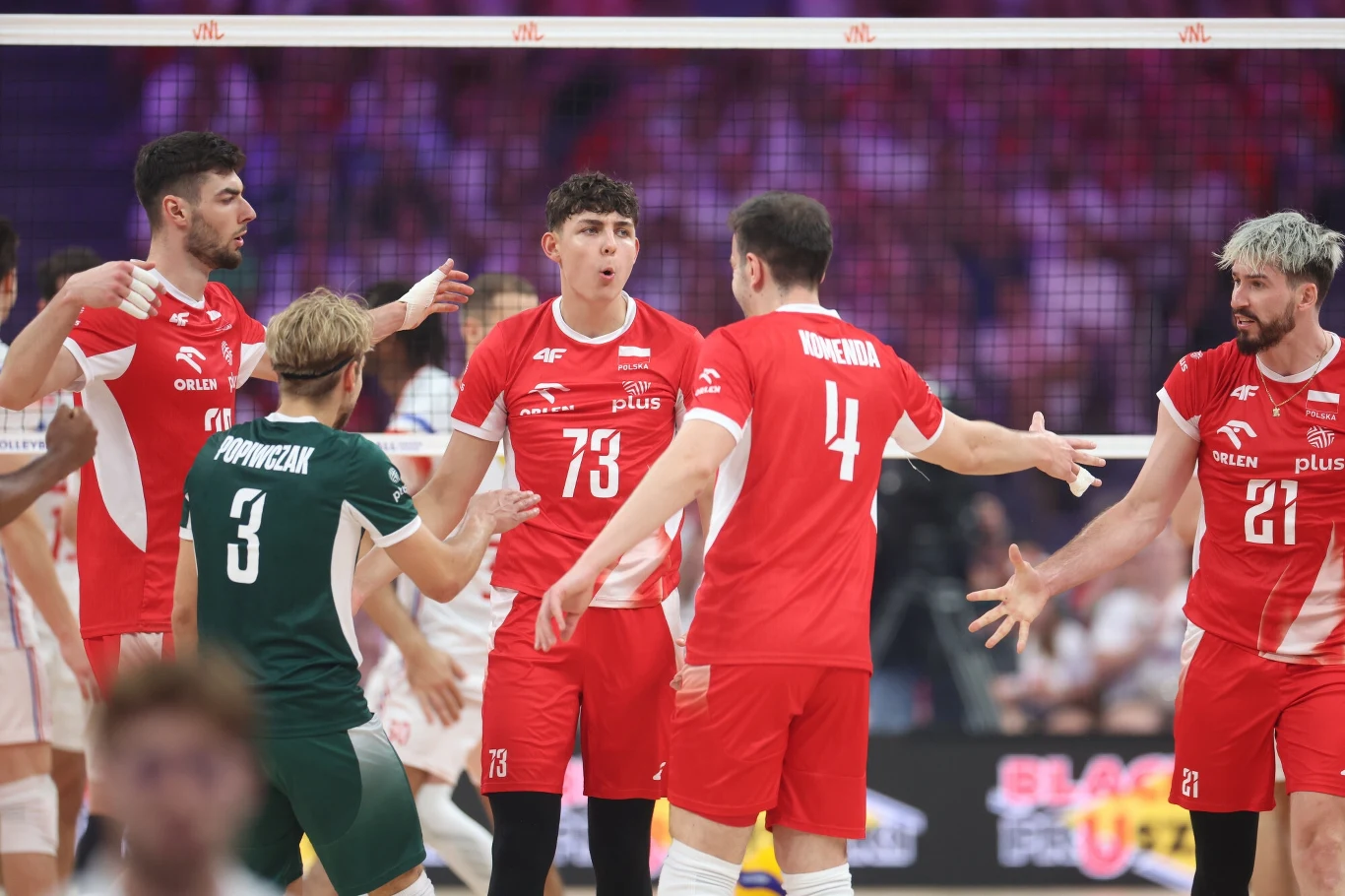 Polscy siatkarze Zawodnicy drużyny siatkarskiej w czerwonych strojach świętują zdobycie punktu na boisku, w tle widoczna siatka oraz rozmyte sylwetki publiczności.