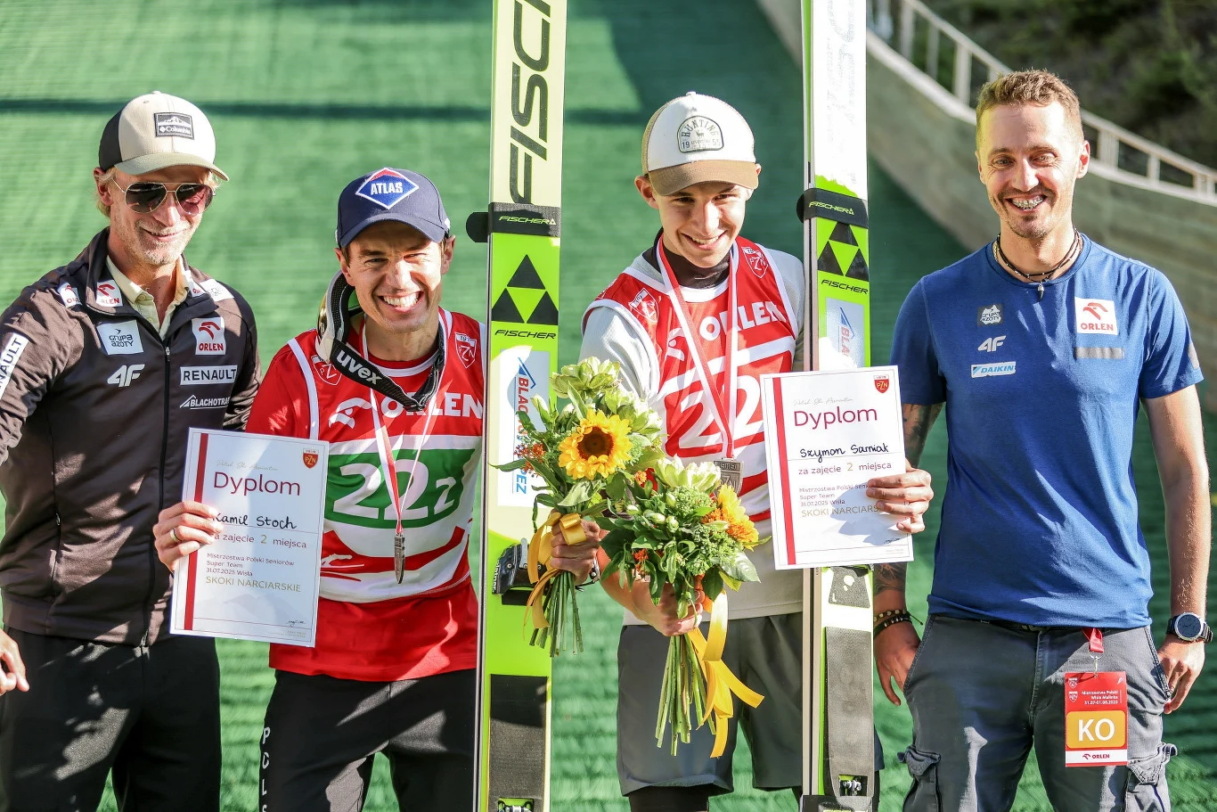 Czterech mężczyzn stojących na tle skoczni narciarskiej, dwóch trzyma dyplomy, jeden bukiet kwiatów, wszyscy uśmiechnięci i ubrani w sportowe stroje, obok nich widoczne narty.