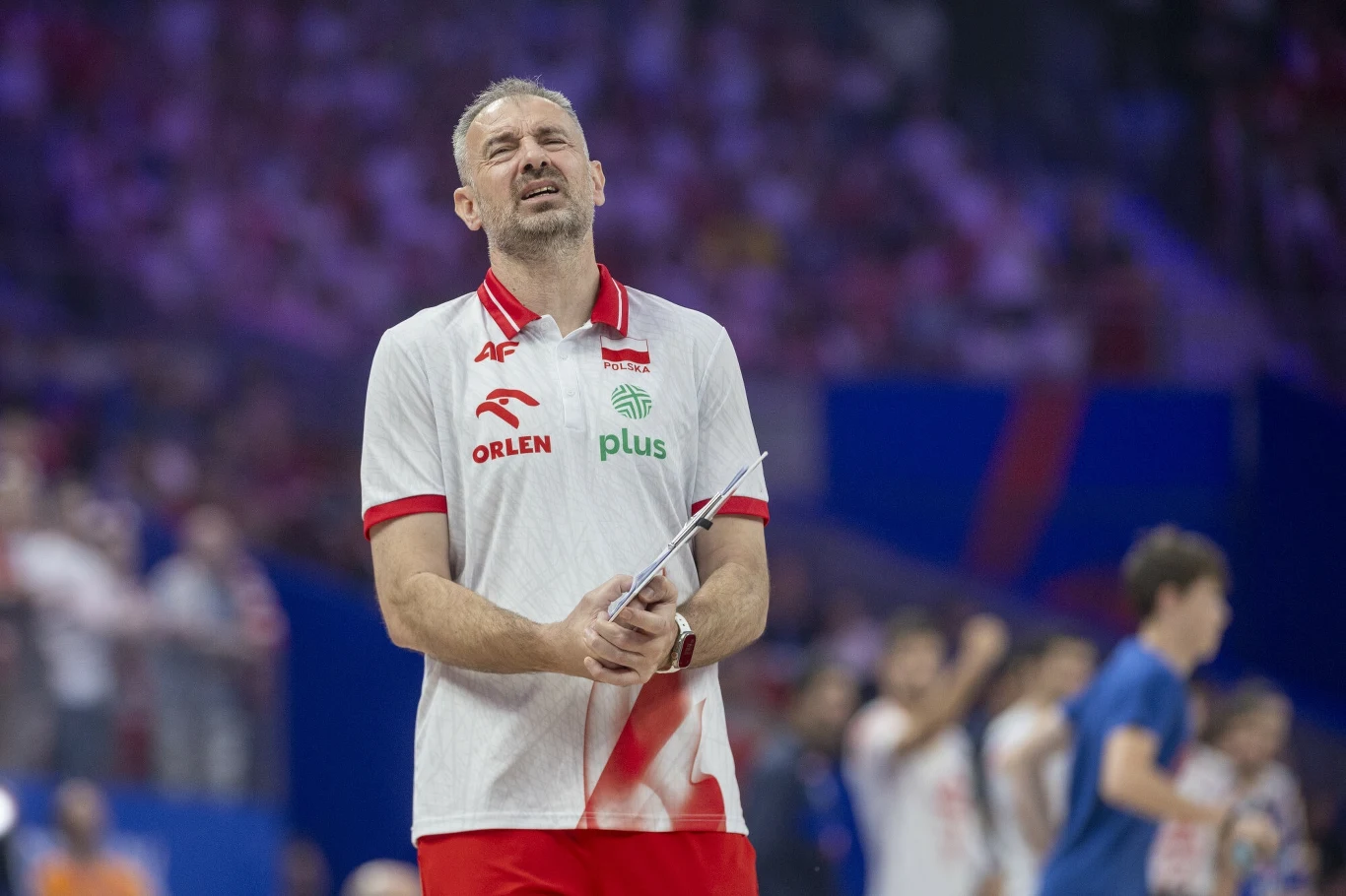 Trener reprezentacji siatkówki w białej koszulce z orzełkiem i logo sponsora trzyma w ręku kartkę, wyrażając silne emocje na tle rozmazanej widowni oraz zawodników.