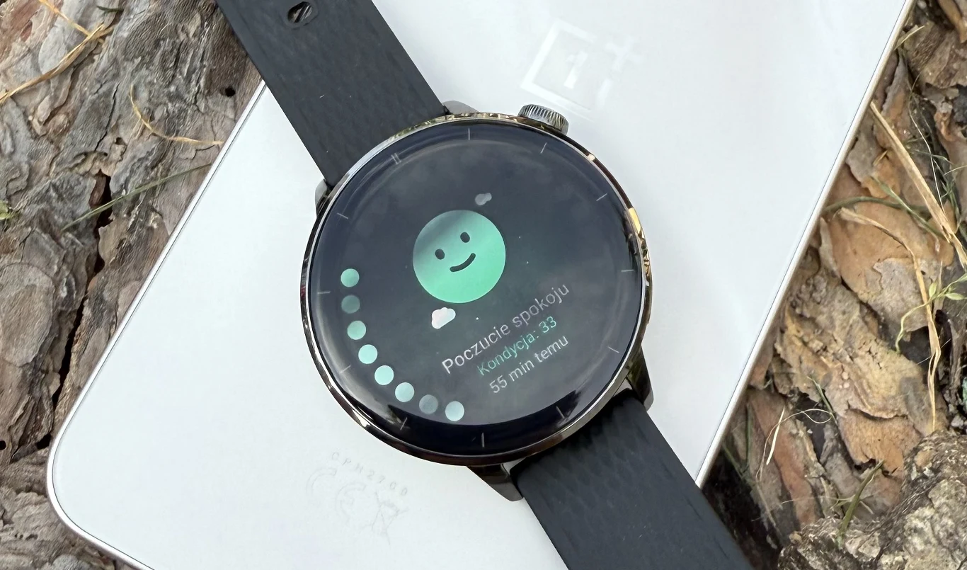 OnePlus Watch 3 43 mm. Funkcjonalny smartwatch w kompaktowym rozmiarze. Czarny smartwatch z okrągłym wyświetlaczem prezentującym ekran dotyczący poczucia spokoju, z uśmiechniętą ikoną i statusem relaksu, leży na białym smartfonie marki OnePlus na tle kory drzewa.