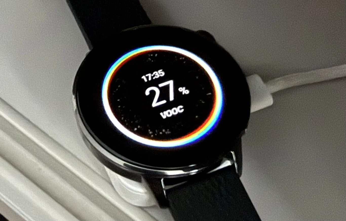 OnePlus Watch 3 wspiera szybkie ładowanie VOOC z użyciem ładowarki o mocy 10 W. Czarny, okrągły smartwatch z paskiem ładuje się poprzez kabel podłączony do jego boku, wyświetlacz pokazuje godzinę 17:35 oraz poziom naładowania baterii wynoszący 27 procent.
