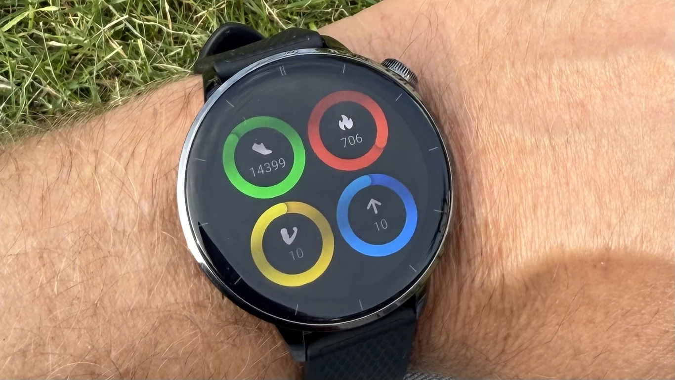 OnePlus Watch 3 to funkcjonalny smartwatch, który zadba o kondycję użytkownika i nie tylko. Czarny smartwatch na nadgarstku wyświetlający okrągły ekran z czterema kolorowymi okręgami, pokazującymi liczbę kroków, spalone kalorie oraz dwie inne statystyki.