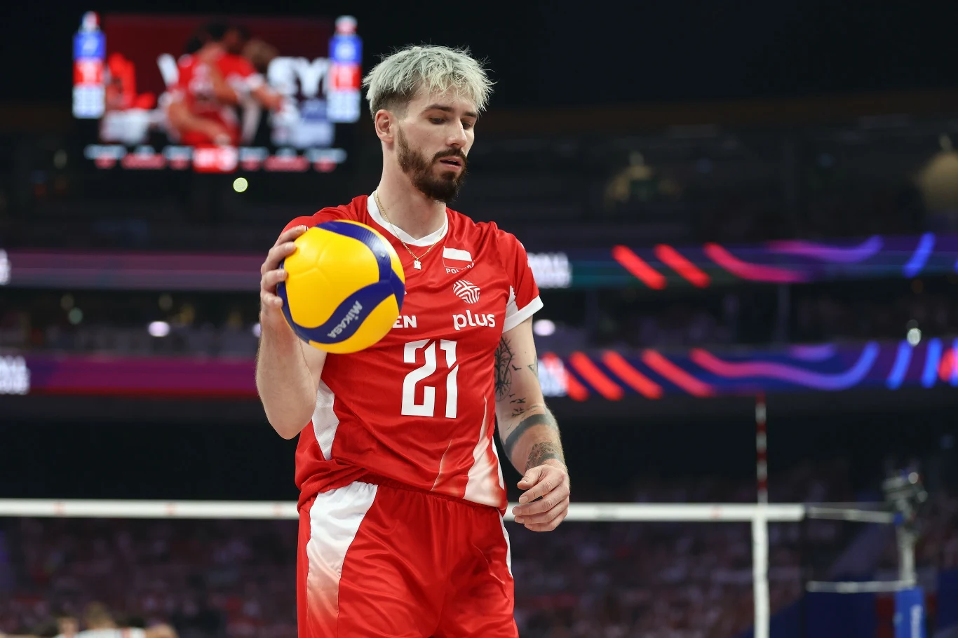 Siatkarz w czerwonym stroju sportowym z numerem 21 przygotowuje się do zagrywki, trzymając piłkę do siatkówki w ręce, w tle niewyraźna publiczność i światła hali sportowej.