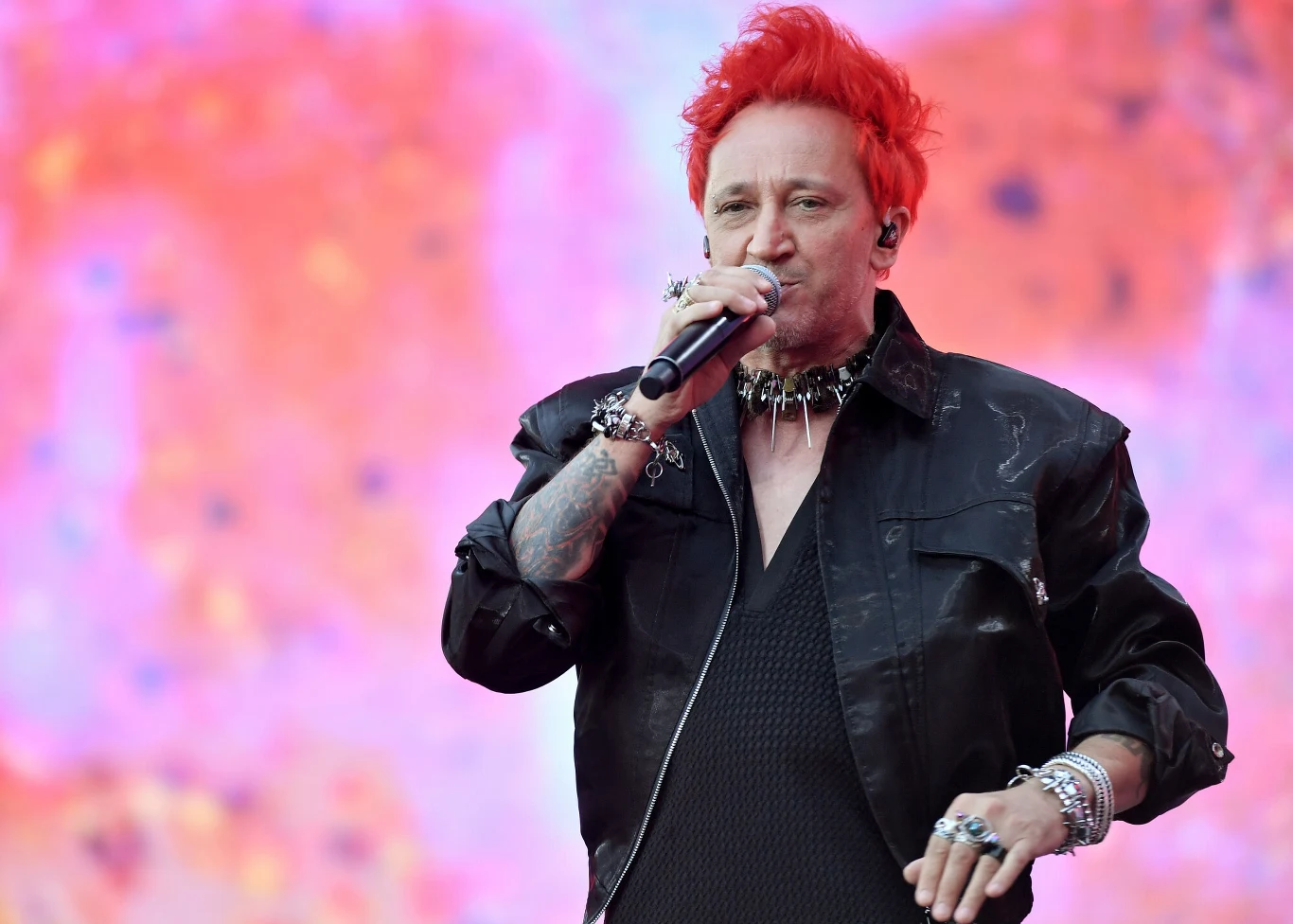 Michał Wiśniewski jest wokalistą Ich Troje Osoba o czerwonych włosach śpiewająca do mikrofonu podczas koncertu, ubrana w czarną kurtkę i liczne bransolety, na zamazanym różowym tle sceny.