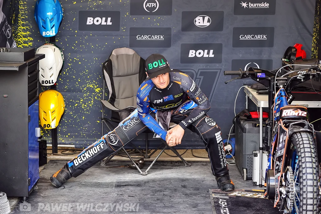 Zawodnik żużlowy rozciąga się na krześle w boksie serwisowym, w otoczeniu kasków ochronnych i motocykla żużlowego przygotowanego do wyścigu.