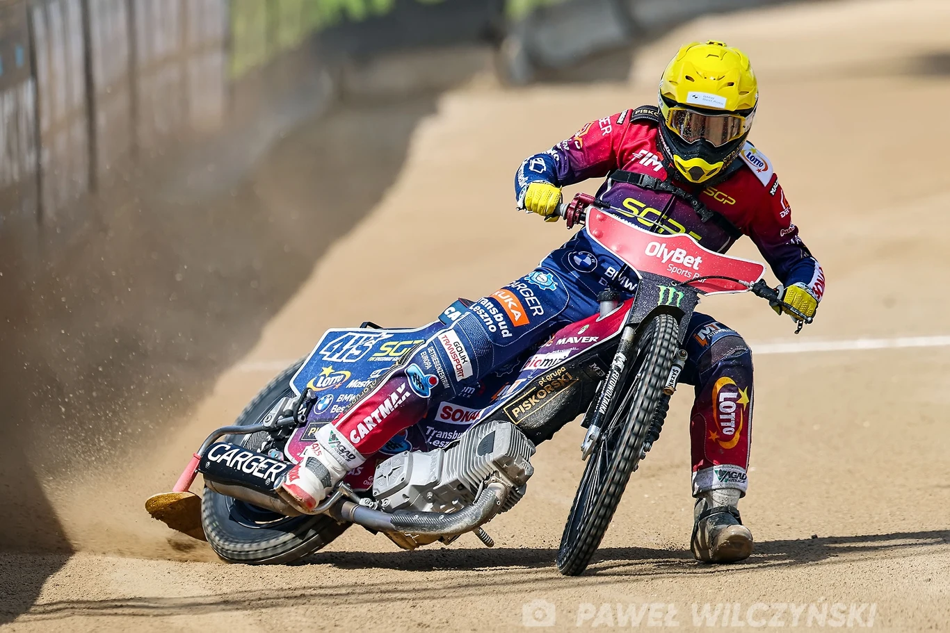 Zawodnik żużlowy ubrany w bogato oznakowany kombinezon sportowy i kask na głowie prowadzi motocykl po torze żużlowym, ostro wchodząc w zakręt, z piaskiem unoszącym się spod tylnego koła.