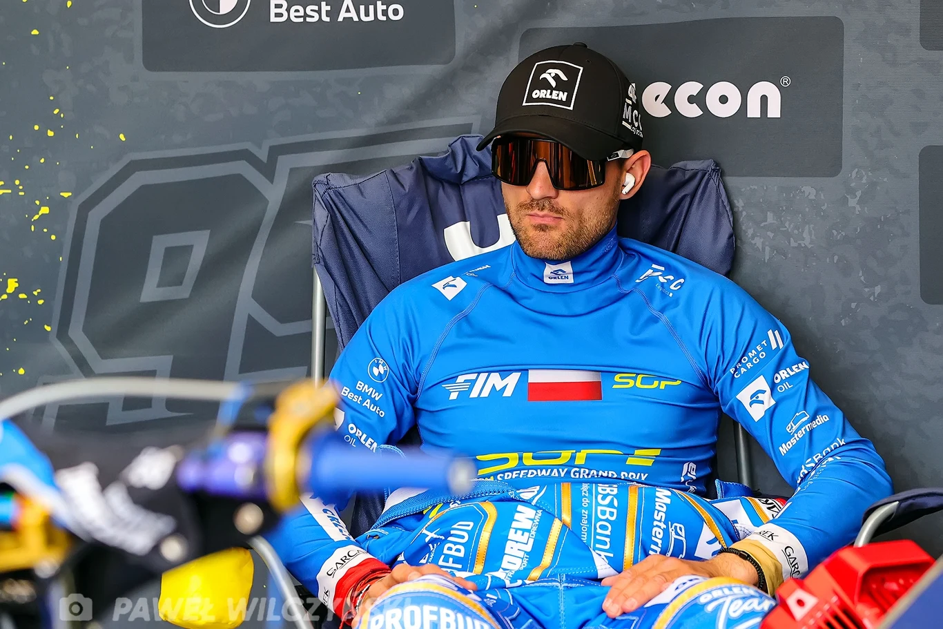 Bartosz Zmarzlik Zawodnik sportów motorowych ubrany w niebieski kombinezon z logo sponsorów, siedzący na krześle w strefie serwisowej, nosi okulary przeciwsłoneczne i czapkę z daszkiem, w tle widoczne tablice reklamowe