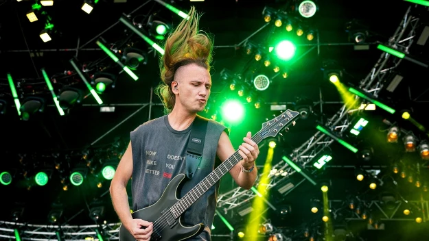 Młody gitarzysta grający na elektrycznej gitarze na scenie koncertowej, dynamiczne oświetlenie z zielonymi reflektorami i ruch w powiewających włosach muzyka podkreślają energię występu.