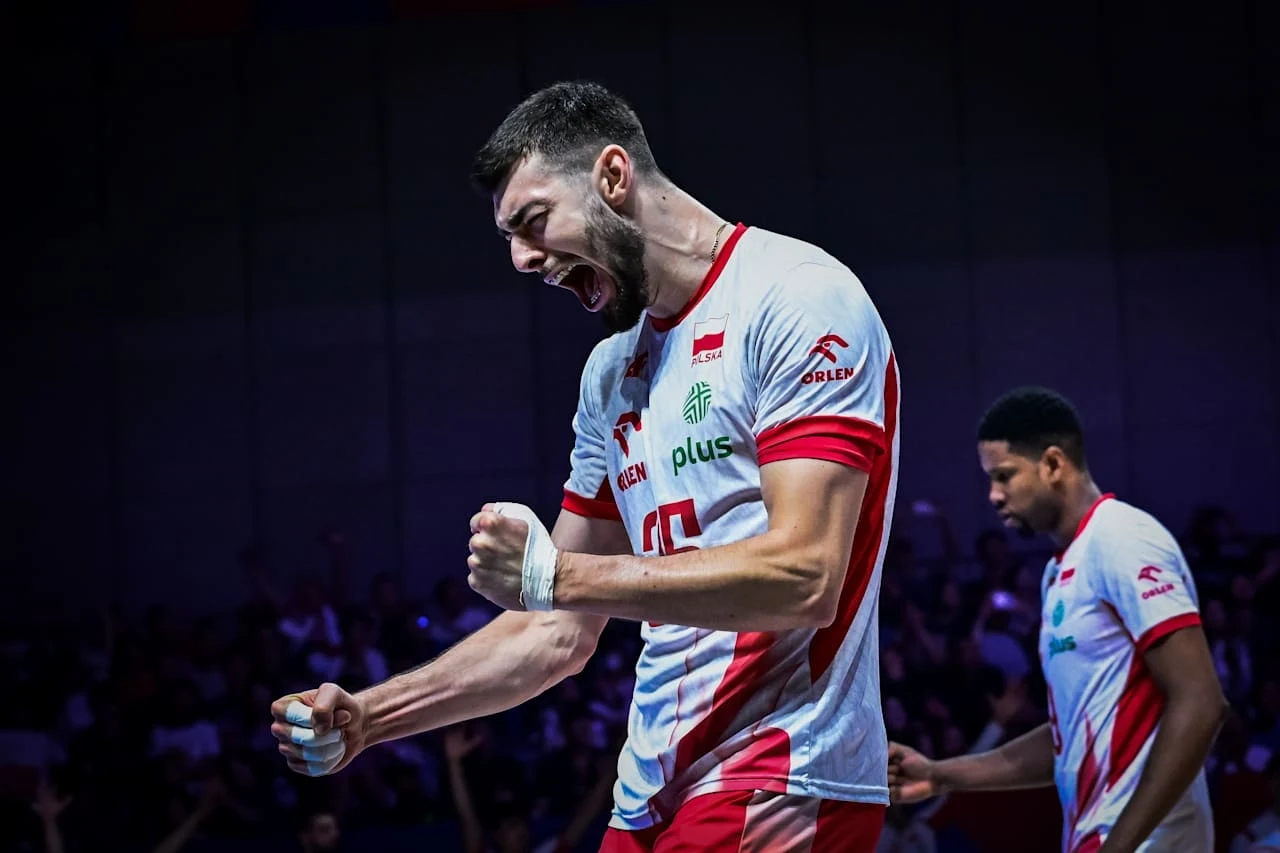 Kewin Sasak Siatkarz w biało-czerwonym stroju drużyny narodowej wyraża silne emocje, zaciskając pięść i krzycząc, w tle znajduje się inny zawodnik oraz rozmyte trybuny pełne kibiców.