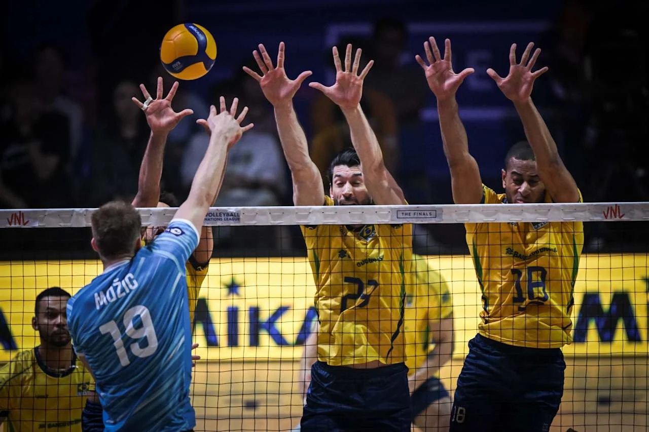 Trzech siatkarzy w żółtych koszulkach podskakuje przy siatce z wyciągniętymi rękami do bloku, naprzeciwko nich znajduje się zawodnik w niebieskiej koszulce próbujący wykonać atak, nad siatką widoczna piłka siatkowa.