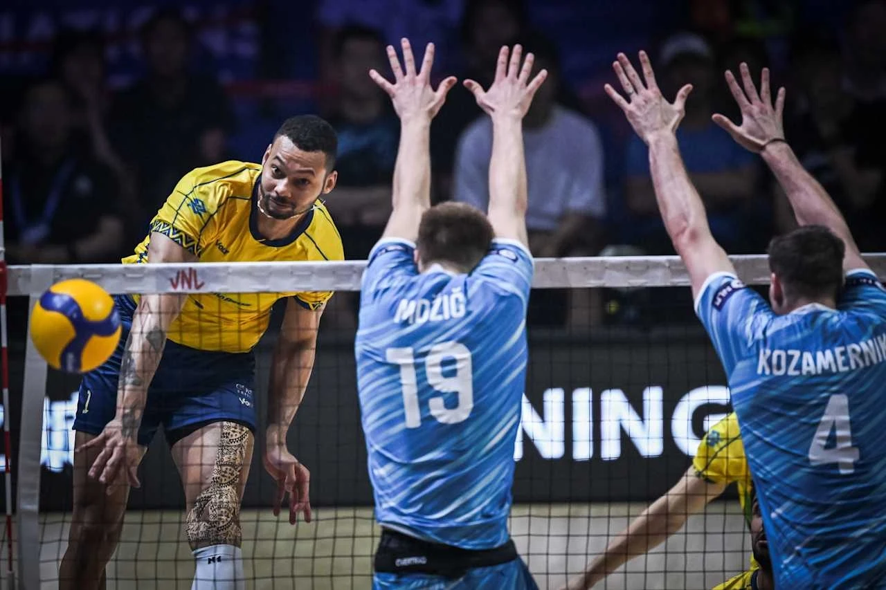 Zawodnik w żółto-niebieskim stroju wykonuje atak siatkarski, a dwóch zawodników w niebieskich koszulkach z numerami 19 i 4 próbuje zablokować piłkę nad siatką podczas meczu siatkówki.