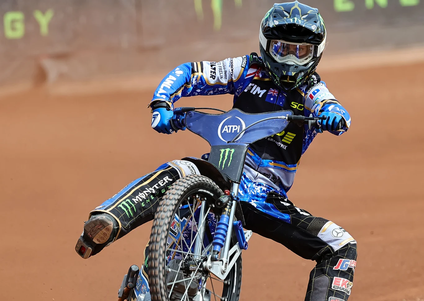 Zawodnik sportów motorowych w profesjonalnym kombinezonie ściga się na motocyklu po torze żużlowym. Wyraziste barwy stroju, dynamiczna poza oraz charakterystyczny pył unoszący się z toru podkreślają sportową atmosferę rywalizacji.