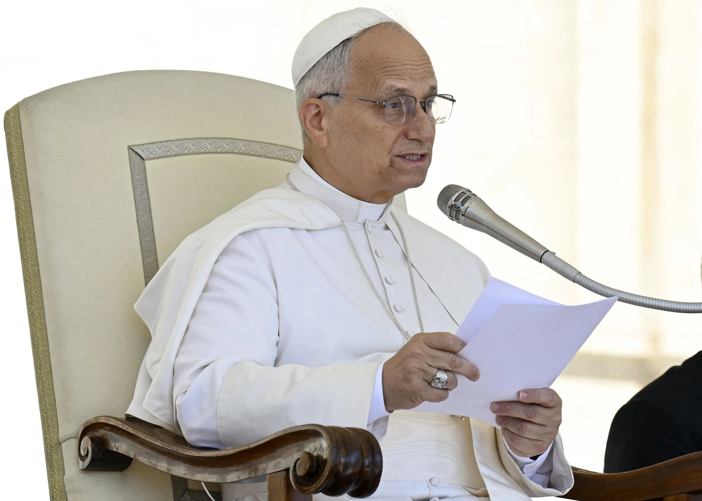 Papież Leon XIV Mężczyzna w białej papieskiej szacie i białej piusce siedzi na dużym fotelu, przemawia do mikrofonu trzymając w ręku kartkę papieru.