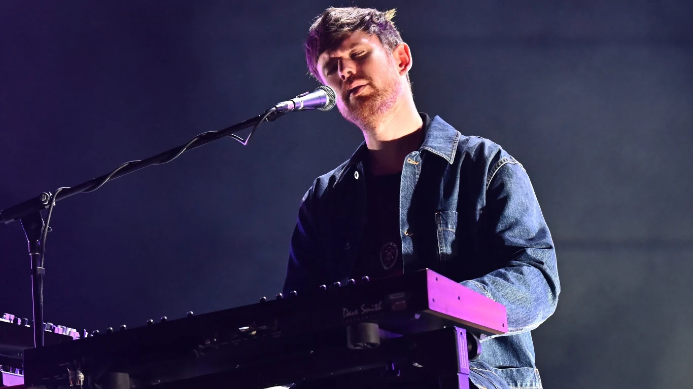 James Blake Mężczyzna grający na syntezatorze i śpiewający do mikrofonu, scena oświetlona światłem koncertowym, artysta ubrany w dżinsową kurtkę na ciemnym tle.