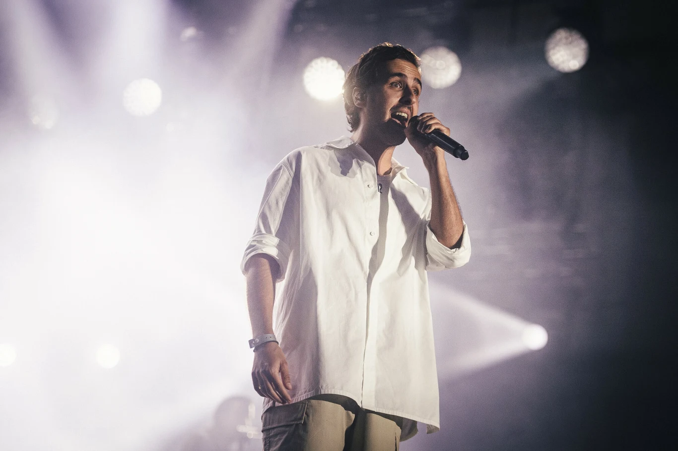 Igo podczas koncertu na Męskim Graniu w Gdańsku Młody mężczyzna w białej koszuli śpiewa do mikrofonu podczas występu na scenie, w tle rozświetlone reflektory i dym nadający koncertowy klimat.