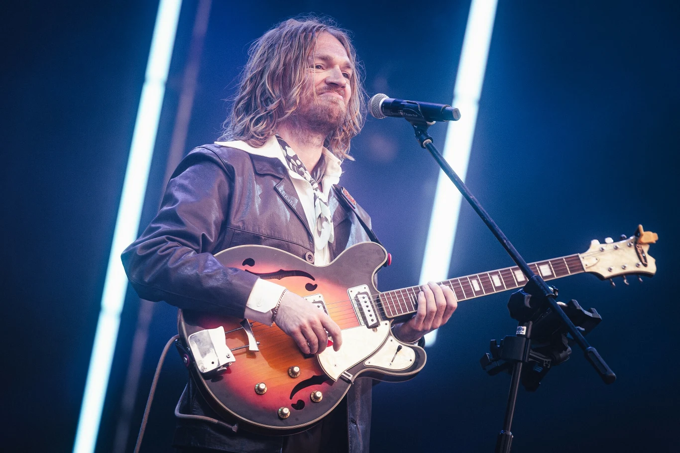 Koncert Cinnamon Gum w Gdańsku na Męskim Graniu 2025 Mężczyzna z długimi włosami w skórzanej kurtce trzyma gitarę elektryczną, stojąc na scenie przy mikrofonie. W tle widoczne są jasne pionowe światła.