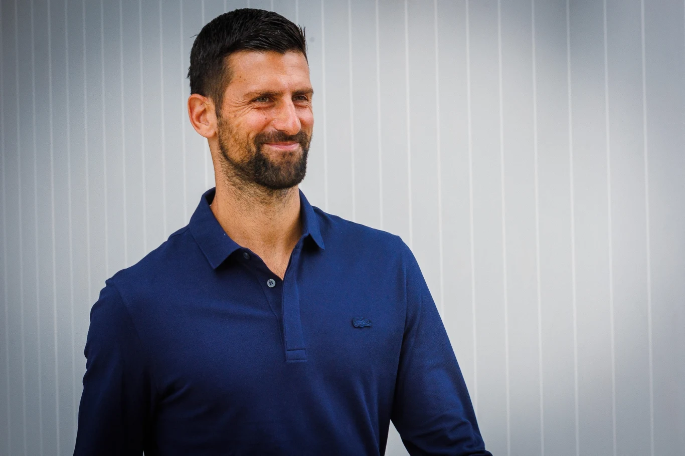 Mężczyzna z krótkimi, ciemnymi włosami i brodą ubrany w ciemnoniebieską koszulkę z kołnierzykiem stoi na tle jasnej, pionowo prążkowanej ściany, uśmiechnięty i patrzący w bok.