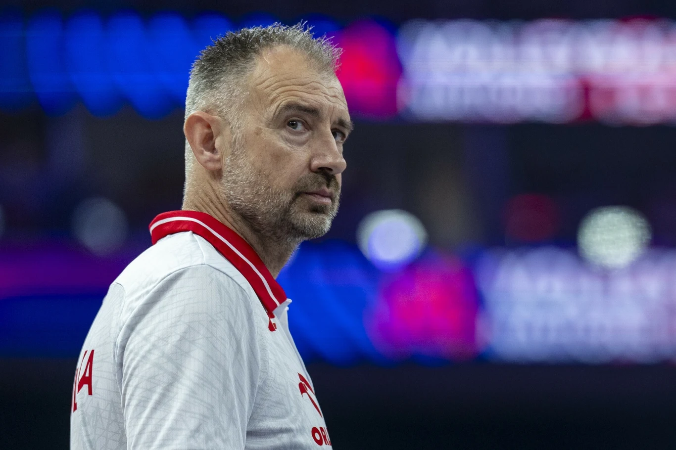 Nikola Grbić Mężczyzna w białej koszulce z czerwonym kołnierzem, z widocznym napisem na plecach, stoi na tle rozmytego, kolorowego tła, wpatrując się w dal.