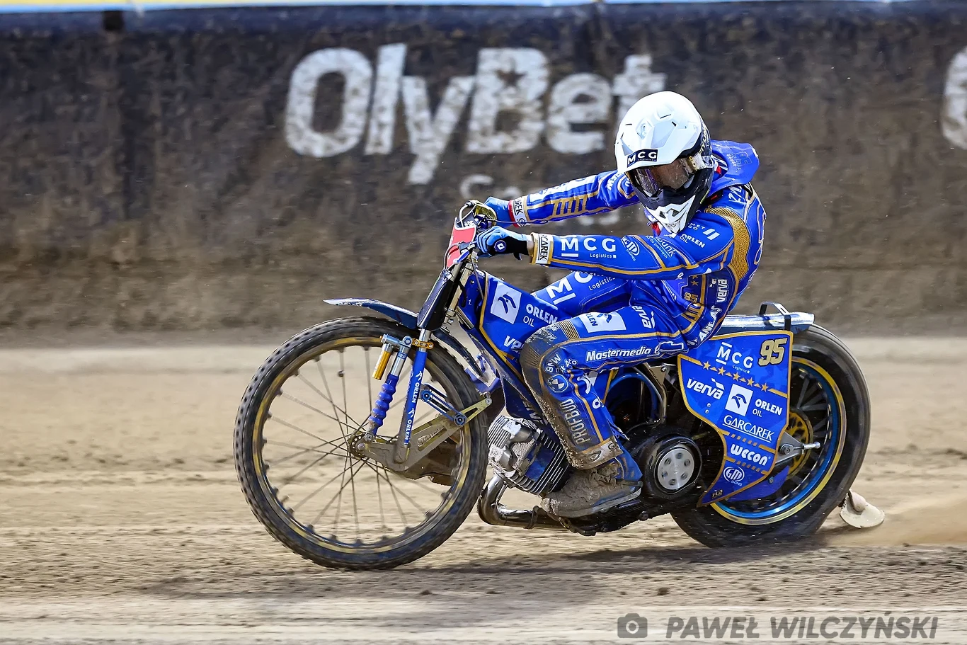 Zawodnik żużlowy w niebieskim kombinezonie i kasku wykonuje dynamiczny zakręt na motocyklu na torze żużlowym, w tle banery reklamowe.