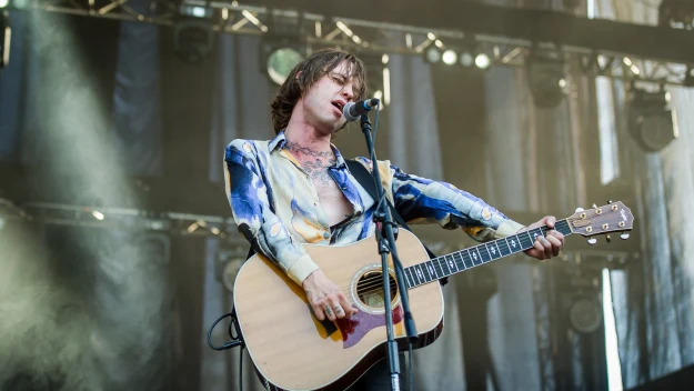 Muzyk w kolorowej koszuli grający na gitarze akustycznej na scenie podczas koncertu, w tle widoczne światła sceniczne i elementy konstrukcji sceny.