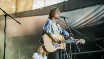 Młody mężczyzna grający na gitarze akustycznej i śpiewający do mikrofonu na scenie podczas koncertu na świeżym powietrzu, w tle widoczny inny członek zespołu.