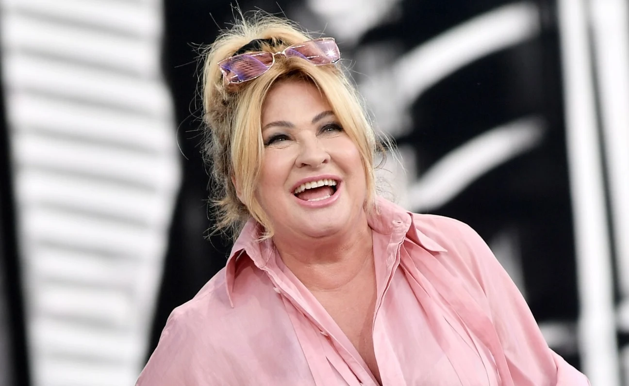 Beata Kozidrak podzieliła się radosną nowiną z fanami Kobieta w średnim wieku z jasnymi włosami spiętymi w niedbały kok, ubrana w różową koszulę, uśmiechnięta, z okularami przeciwsłonecznymi na głowie, na tle rozmytego czarno-białego wzoru.