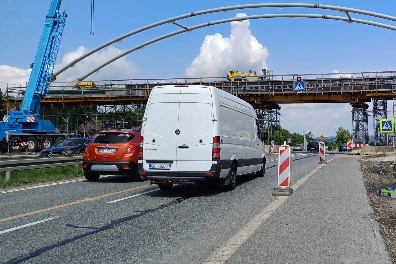 Od 4 sierpnia kierowcy muszą przygotować się na utrudnienia w związku z pracami przy budowie węzła Myślenice Ruch samochodowy na drodze w pobliżu budowy wiaduktu, widoczny biały bus i pomarańczowy samochód osobowy, w tle konstrukcja mostu z rusztowaniami oraz niebieski dźwig.