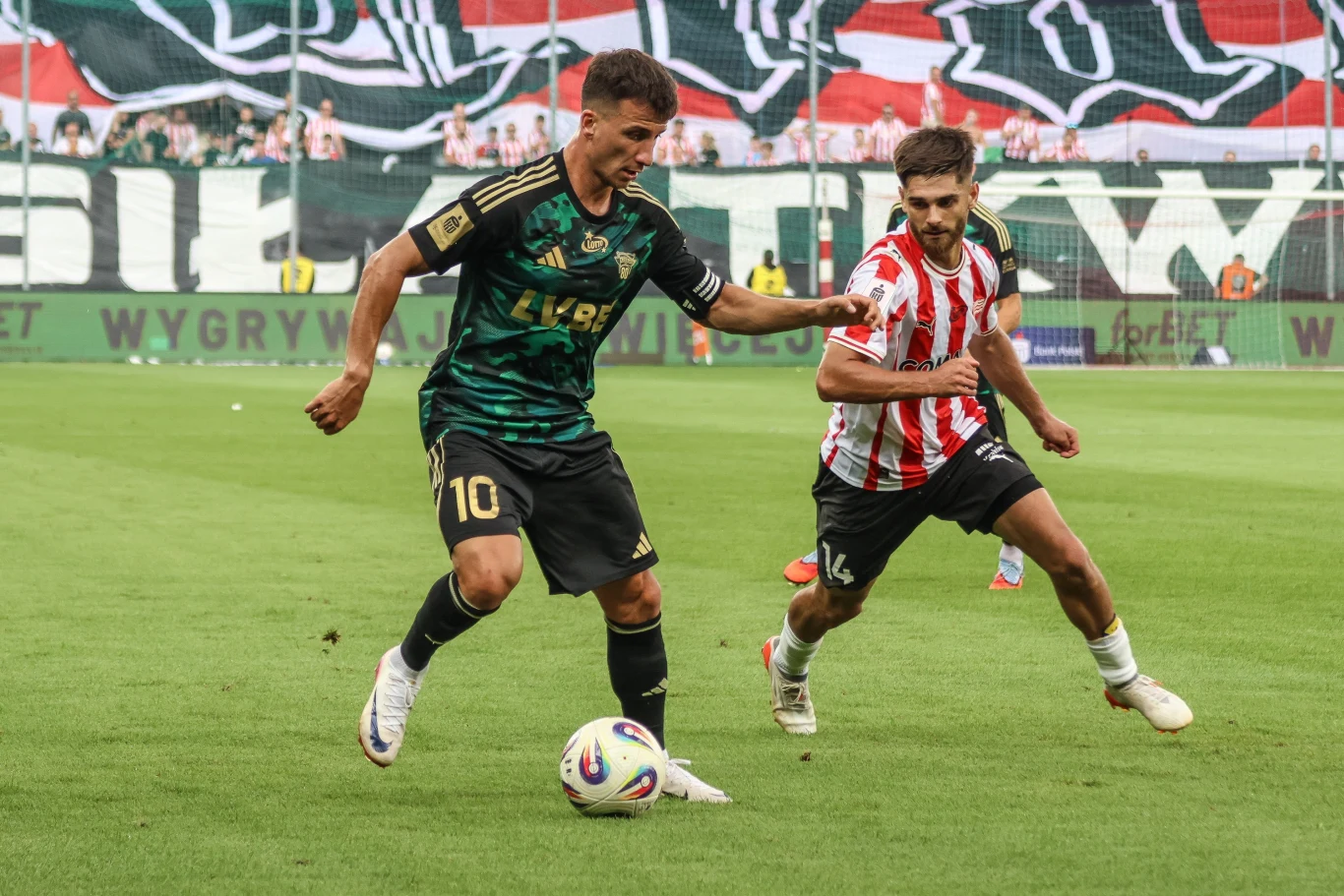 Dwóch piłkarzy podczas meczu piłki nożnej rywalizuje o piłkę na zielonej murawie stadionu, w tle widać trybuny z kolorowym transparentem i kibicami.