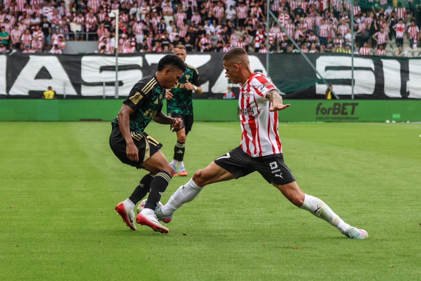 Dwóch piłkarzy walczących o piłkę na zielonym boisku podczas meczu, jeden ubrany w zielono-czarną koszulkę, drugi w biało-czerwone pasy, w tle widoczni kibice na trybunach.