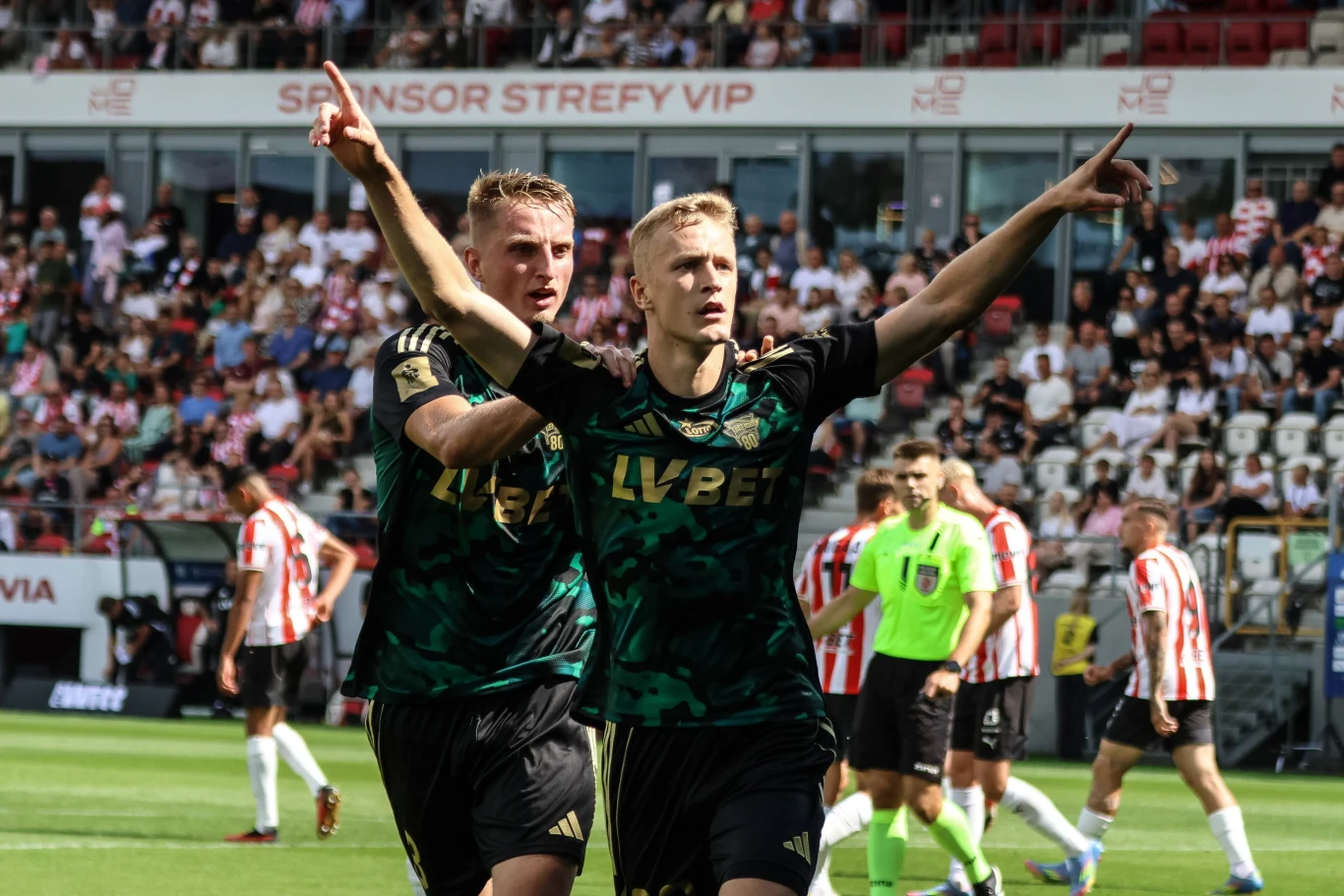 Dwóch piłkarzy w zielonych strojach świętuje zdobycie bramki na boisku, jeden z nich unosi obie ręce w geście zwycięstwa, wokół nich znajdują się inni zawodnicy oraz widzowie na trybunach stadionu.