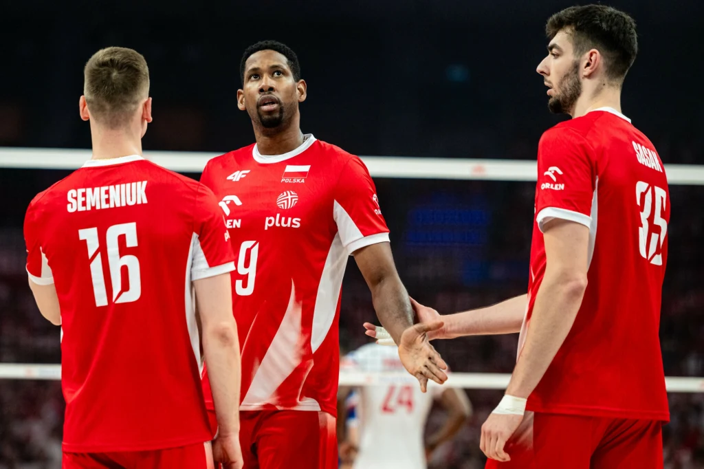 Trzech siatkarzy w czerwonych koszulkach polskiej reprezentacji podczas meczu, jeden z nich wyciąga rękę w kierunku kolegi z drużyny, wyrażając gest wsparcia lub motywacji.