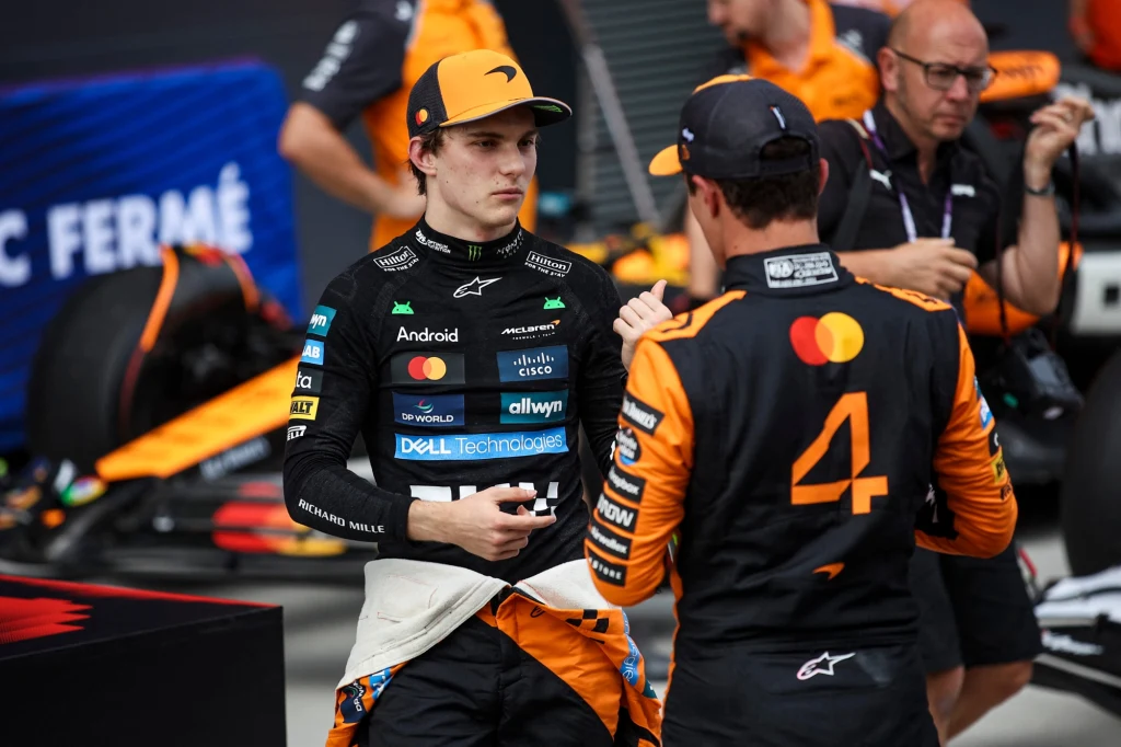 Oscar Piastri w rozmowie z Lando Norrisem Dwóch kierowców wyścigowych w kombinezonach zespołu McLaren rozmawia ze sobą w strefie parku zamkniętego, w tle widoczni członkowie zespołu i fragmenty bolidów wyścigowych.