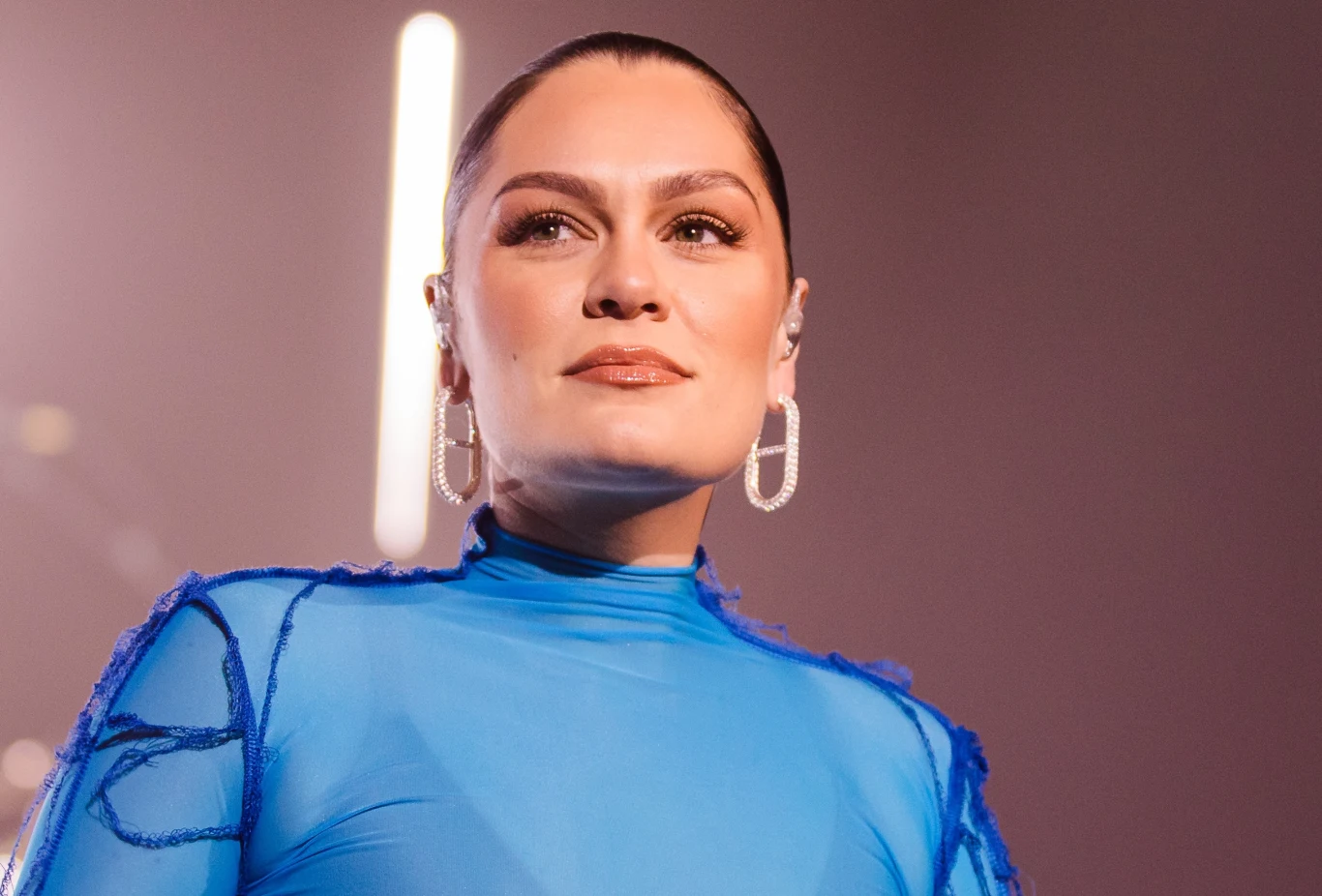 Jessie J ponownie trafiła do szpitala Kobieta w niebieskim, przezroczystym kostiumie sceniczny, z elegancko zaczesanymi do tyłu włosami, nosząca duże, błyszczące kolczyki, na tle jasnego oświetlenia.