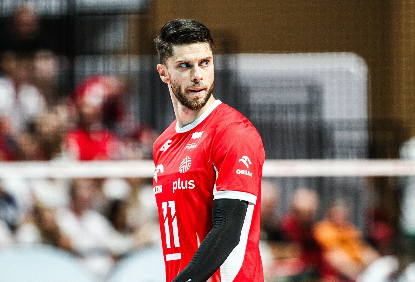 Siatkarz w czerwonej koszulce z numerem 11 skupiony podczas meczu, w tle widoczni kibice i fragment siatki.