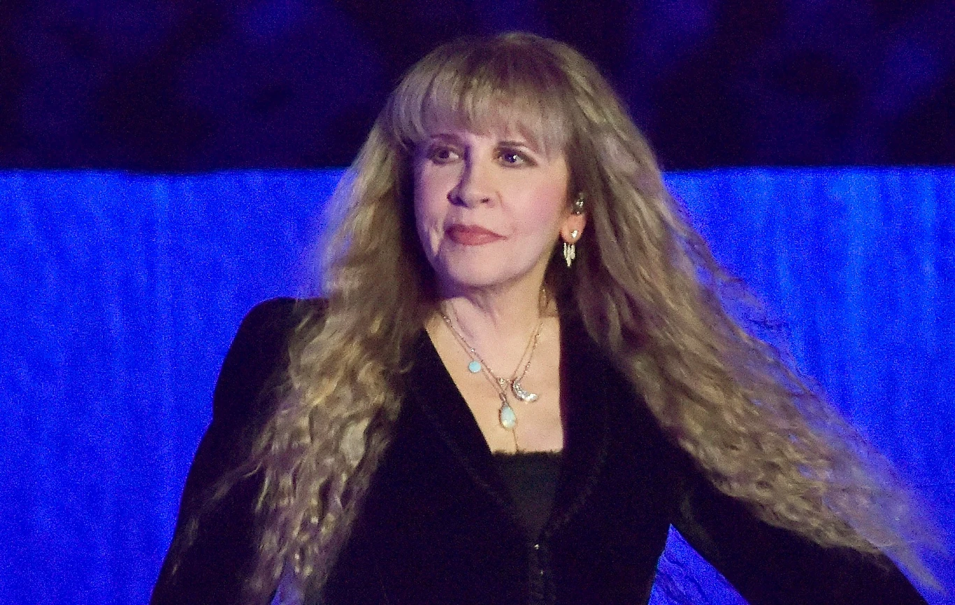 Stevie Nicks przełożyła część swoich planowanych koncertów Kobieta z długimi blond włosami ubrana w czarną marynarkę, ozdobiona delikatną biżuterią, stoi na tle niebieskiego tła wyglądająca pewnie przed siebie.