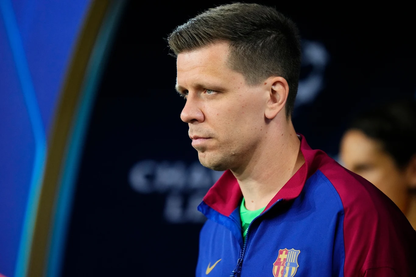 Mężczyzna w kurtce klubowej FC Barcelona z poważnym wyrazem twarzy stoi na tle rozmytego niebieskiego tła z napisem Champions League.