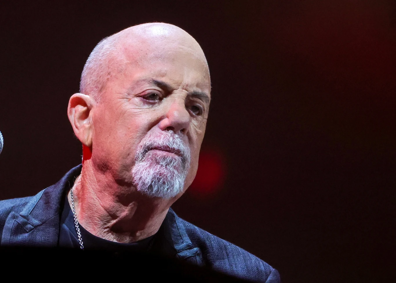 Billy Joel doczekał się dokumentu o swoim życiu Starszy mężczyzna z łysą głową i siwiejącą brodą, ubrany w ciemny garnitur i koszulkę, znajduje się na scenie przy mikrofonie, na tle rozmytego, ciemnego tła z czerwoną poświatą.