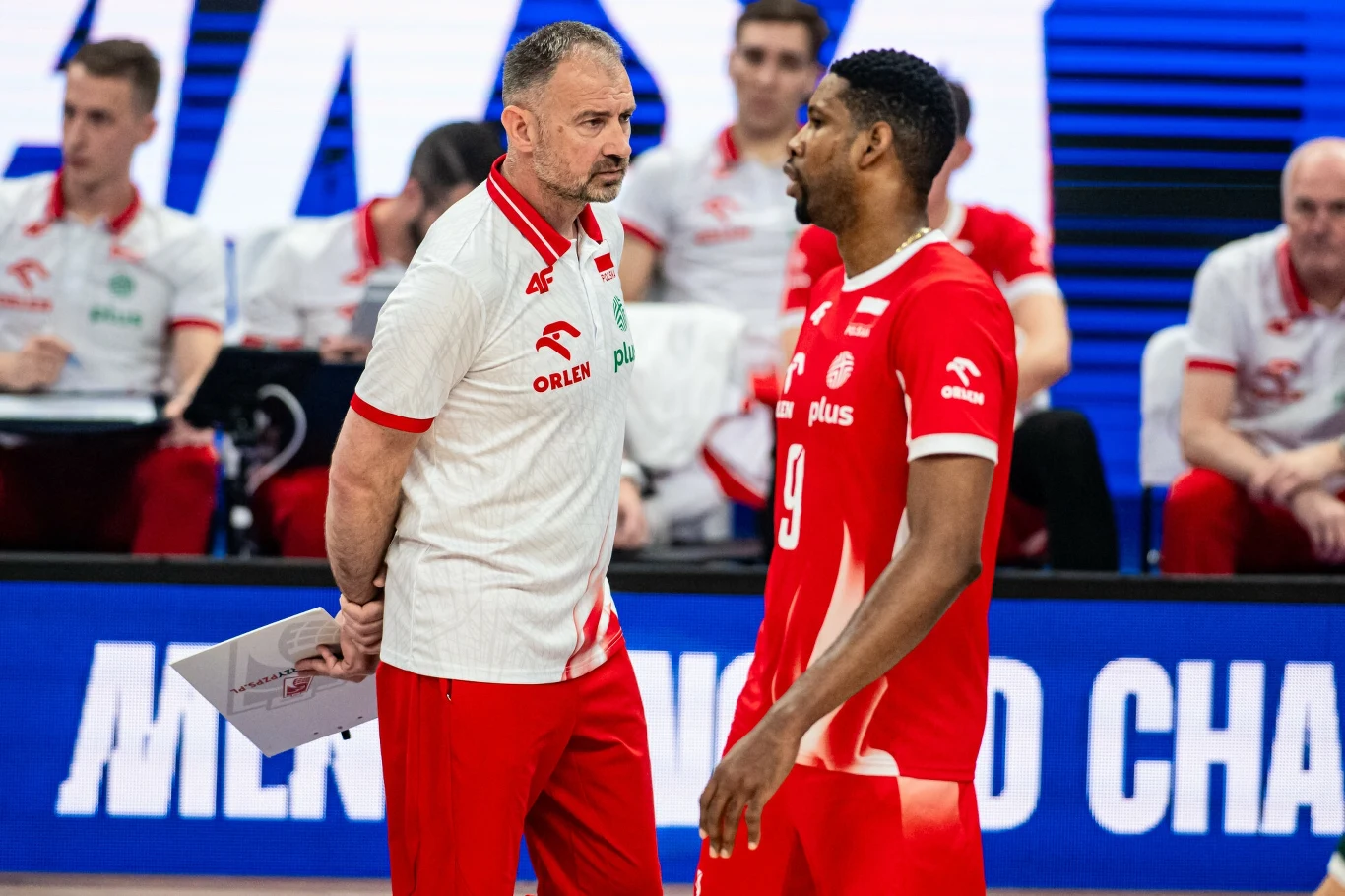Trener oraz zawodnik reprezentacji siatkarskiej Polski podczas rozmowy na meczu, w tle pozostali członkowie sztabu trenerskiego i ekrany LED z napisem turnieju.