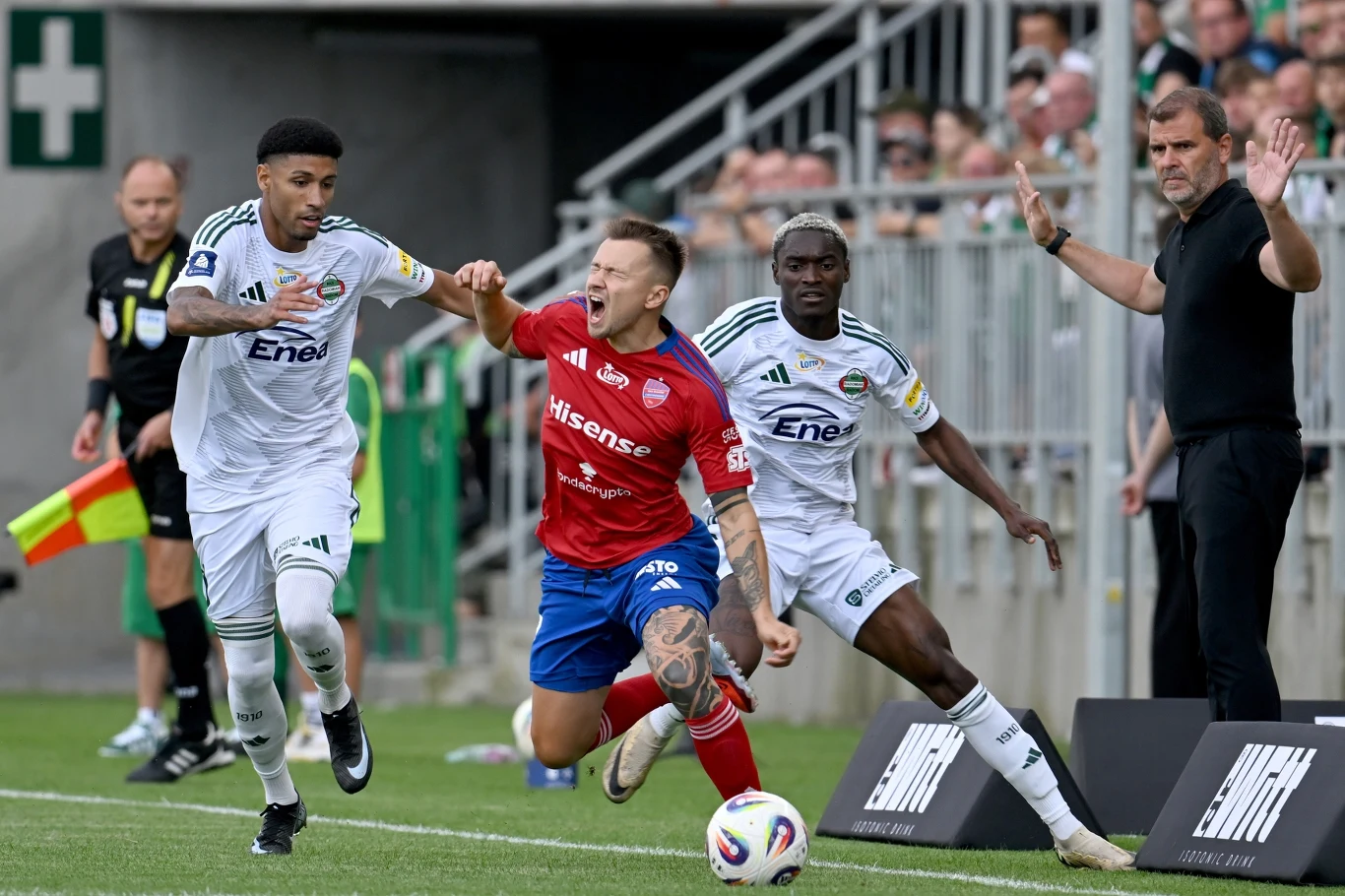 Capita i Fran Tudor Dwóch piłkarzy w białych strojach biegnie obok zawodnika w czerwonej koszulce, który upada podczas dynamicznej akcji na boisku, a sędzia boczny oraz trener obserwują sytuację z boku.