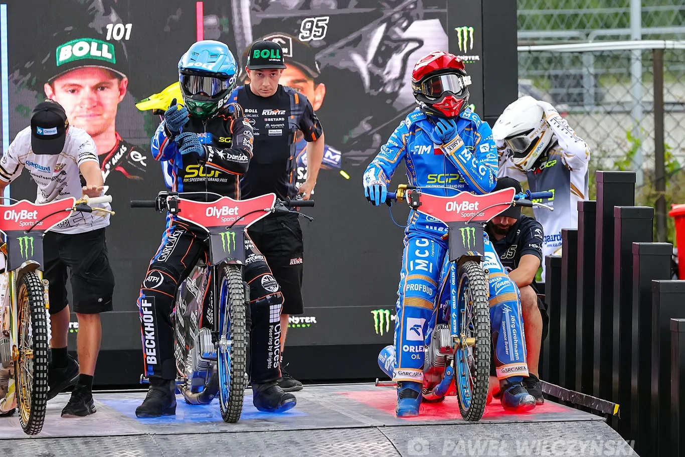 Dwóch żużlowców w kolorowych kombinezonach i kaskach stoi na motocyklach gotowych do startu, obok nich mechanicy dokonują ostatnich regulacji, w tle banery sponsorów oraz duże zdjęcia innych zawodników.