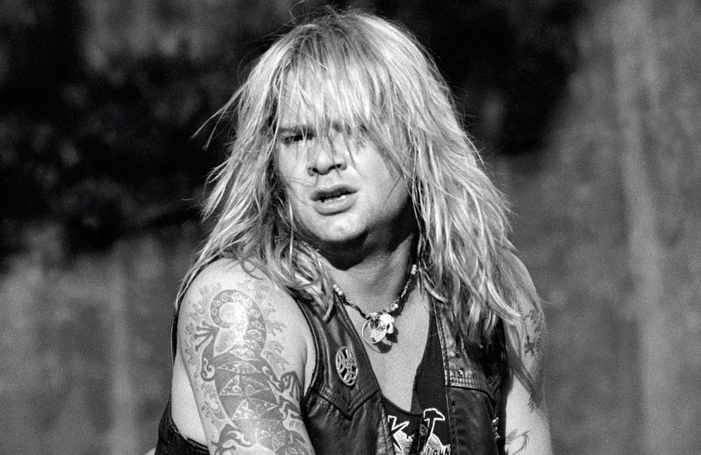 Długowłosy mężczyzna z licznymi tatuażami na ramieniu, ubrany w skórzaną kamizelkę i naszyjnik z zawieszką, patrzący przed siebie z dynamicznym wyrazem twarzy.